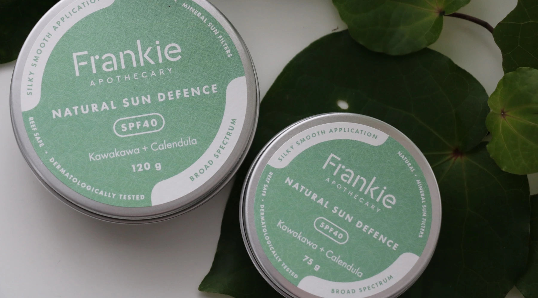 Frankie Apothecary Natural Sun Defence SPF 40 tins featuring Kawakawa and Calendula, placed on fresh green leaves at Intentionally Natural NZ.