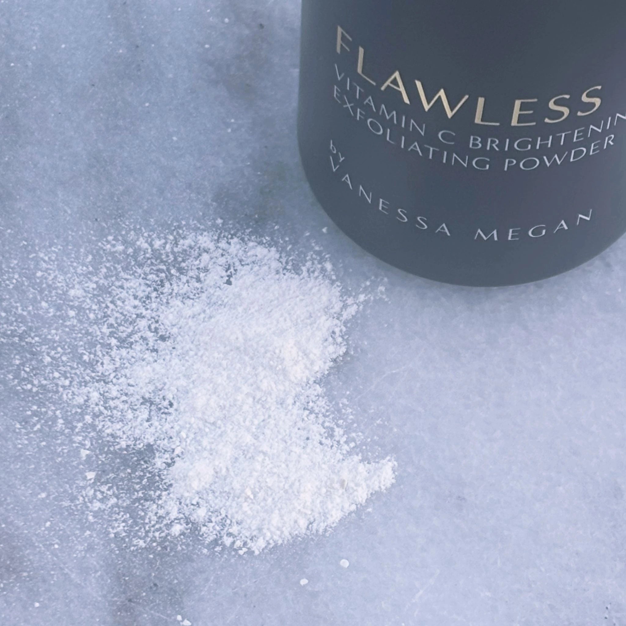 Close-up of the Metamorphosis Flawless Exfoliating Powder scattered on a light marble surface, showcasing its fine, powder texture beside the bottle.