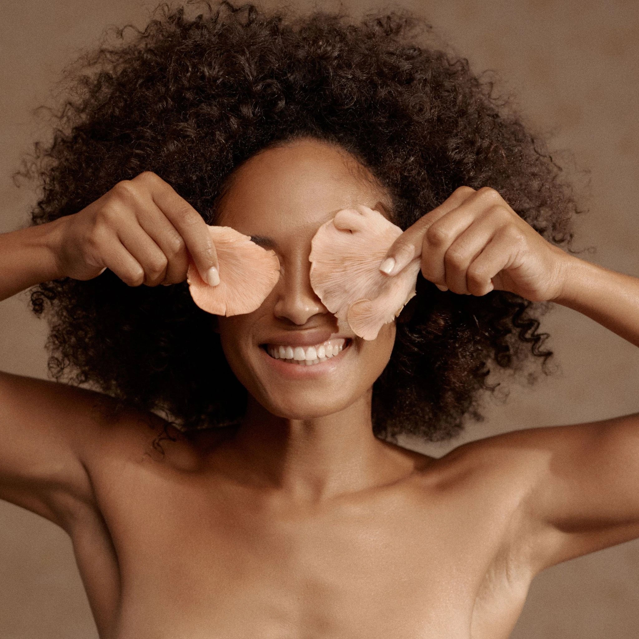 Smiling woman playfully holding pink oyster mushrooms over her eyes.