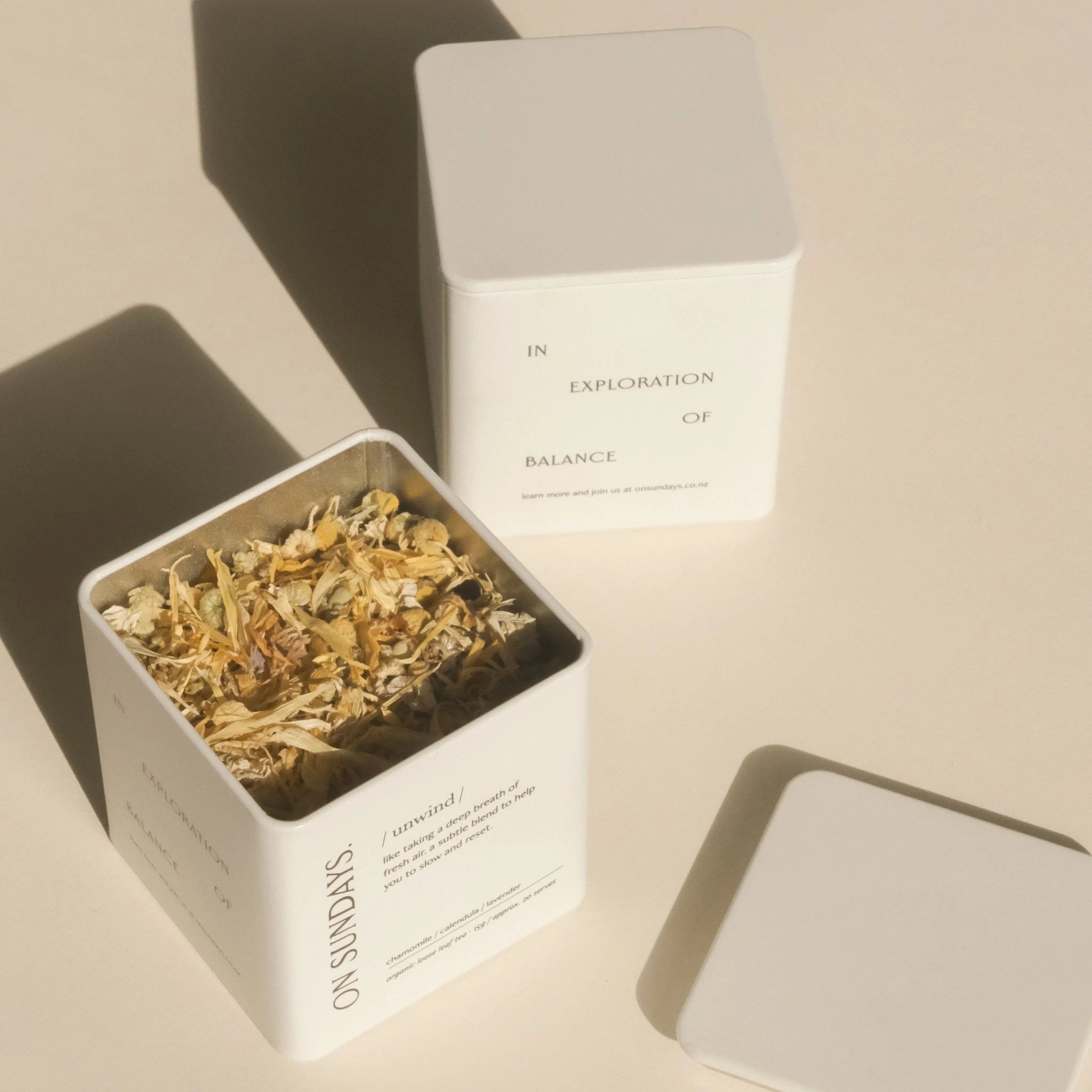 On Sundays Unwind herbal tea in signature white tin with lid, filled with loose organic botanicals including chamomile, calendula, and lavender, shot in soft natural light.