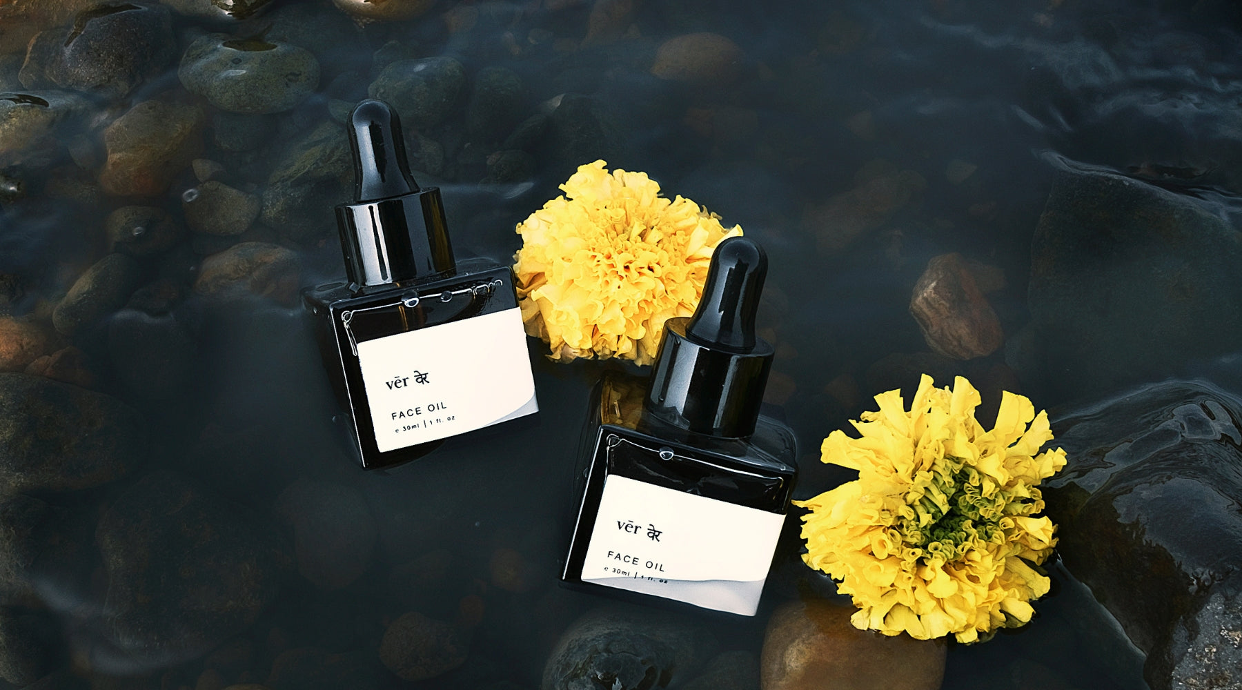 ver The Ritual Face Oil bottles resting on natural river stones in flowing water, surrounded by vibrant yellow flowers.