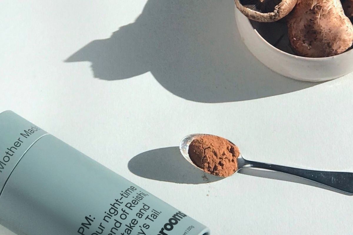 Mother Made PM Mushroom Powder tube beside a spoonful of powder and a bowl of raw mushrooms on a soft grey surface.