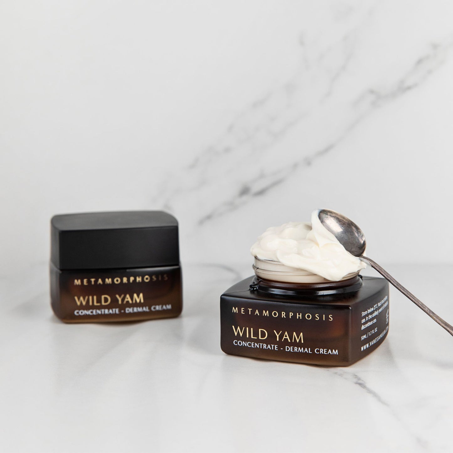 Two jars of Metamorphosis Wild Yam Concentrate - Dermal Cream on a white marble surface, with one jar open showing white cream and a spoon resting on top.