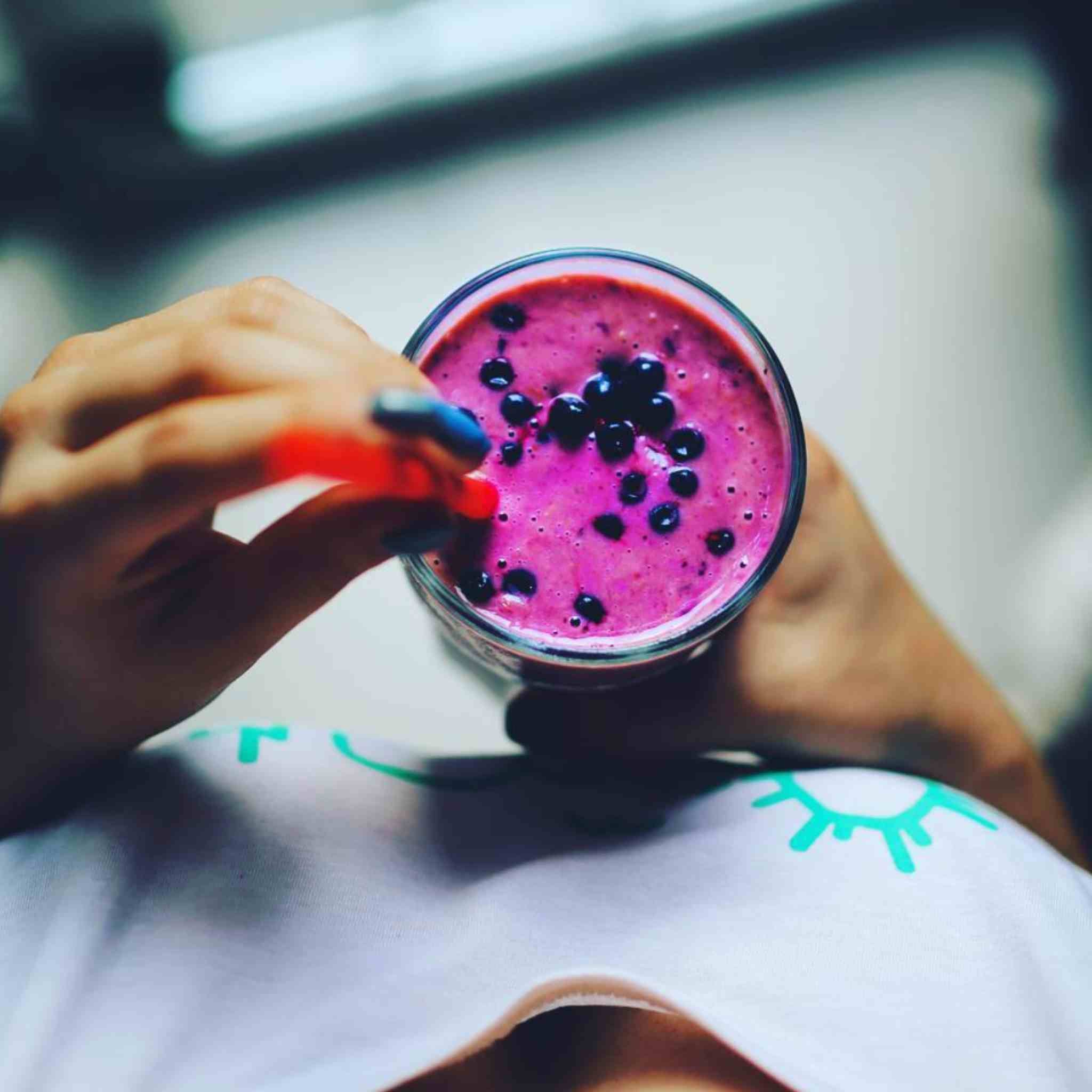 A close-up lifestyle shot of a person holding a glass of vibrant magenta smoothie made with the Matakana Superfoods Organic Maqui Berry Powder topped with fresh blueberries, using a red straw. The person is wearing a white t-shirt with a green graphic print, set against a blurred background.
