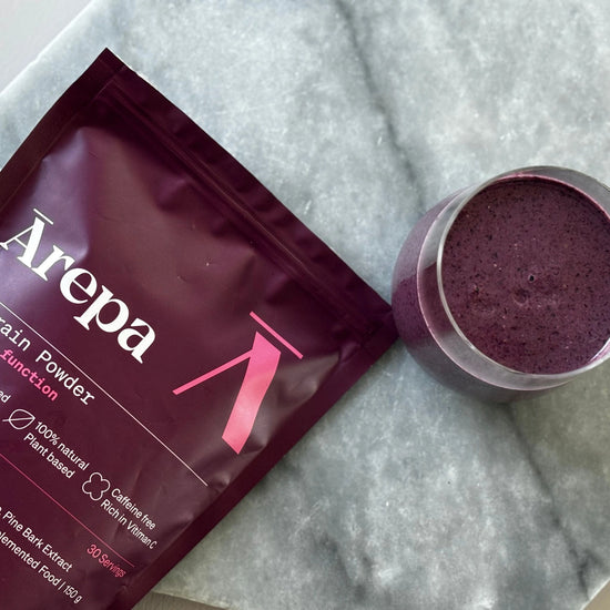 Ārepa Brain Powder pouch next to a glass of blended purple smoothie, showcasing its use in functional beverages for mental clarity and focus.