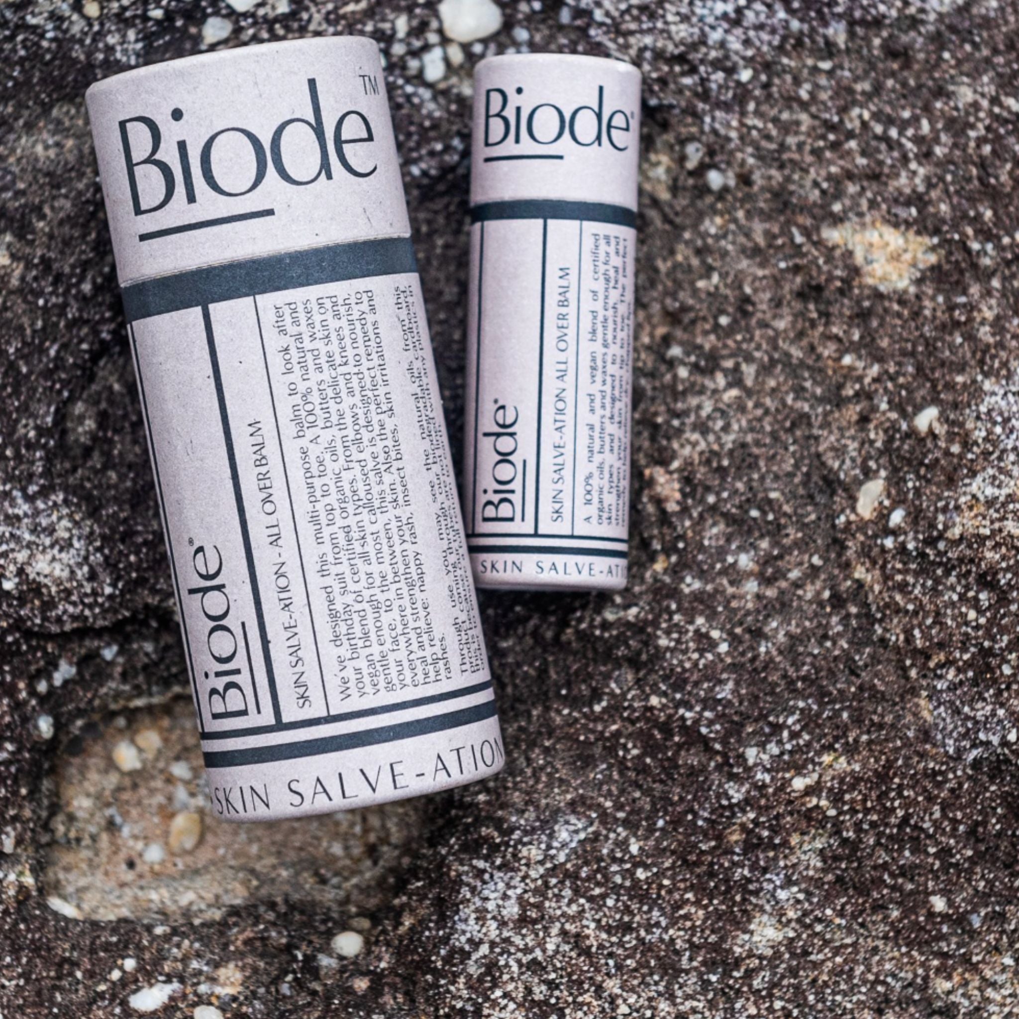 Two Biode skin salve tubes lying side by side on a textured stone surface. Large size 60g on the left and 10g on the right.