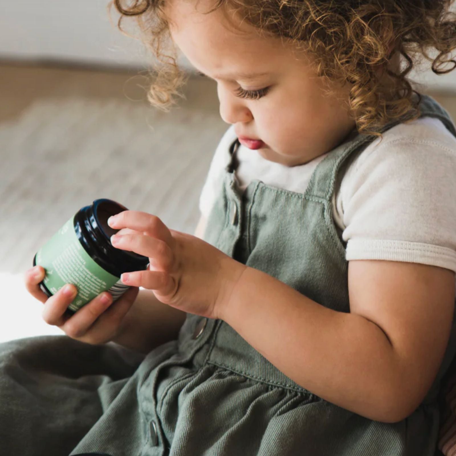 A young child with curly hair looks down at a jar of the Frankie Apothecary Kawakawa Repair Balm held in their hands, wearing a white t-shirt and green denim overalls in a softly lit indoor setting.
