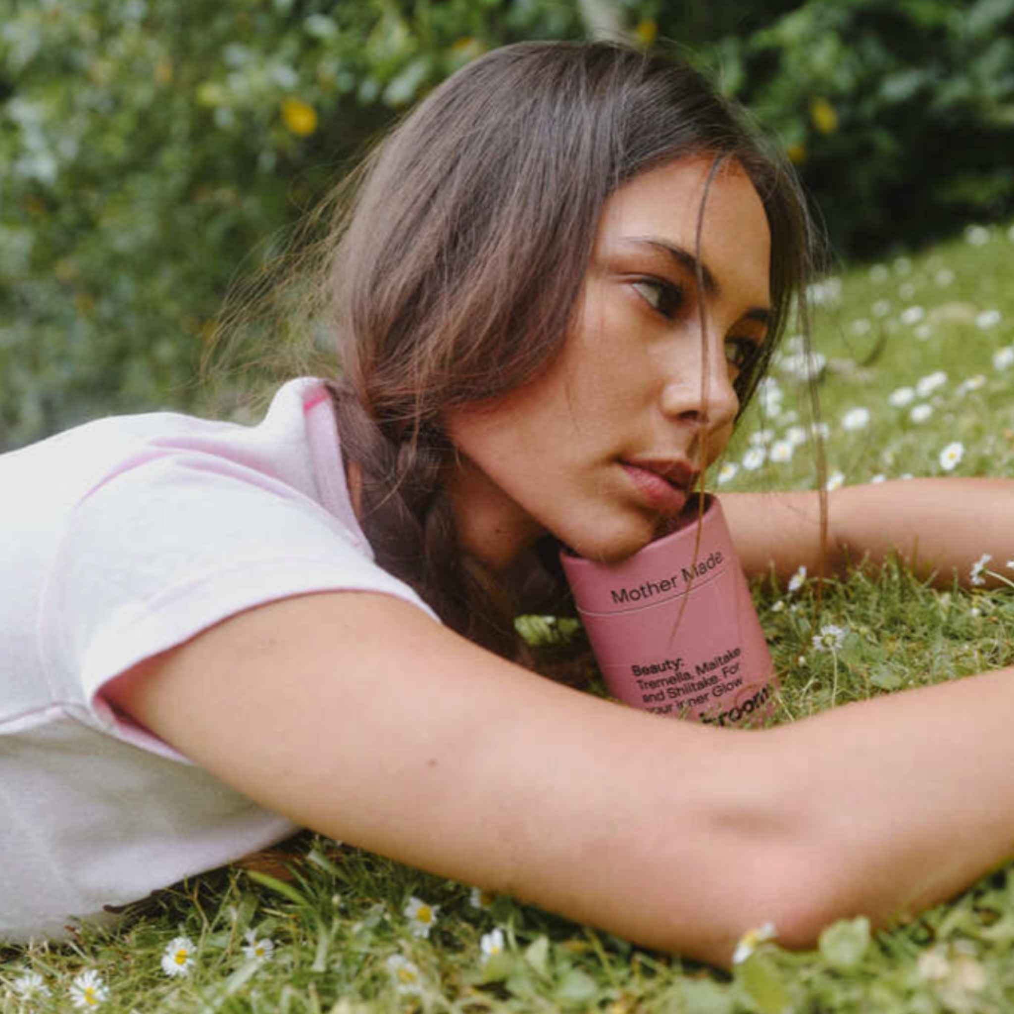 A woman lying on grass outdoors with a pink tube lof the Mother Made  Beauty Blend with a blurred green garden background.