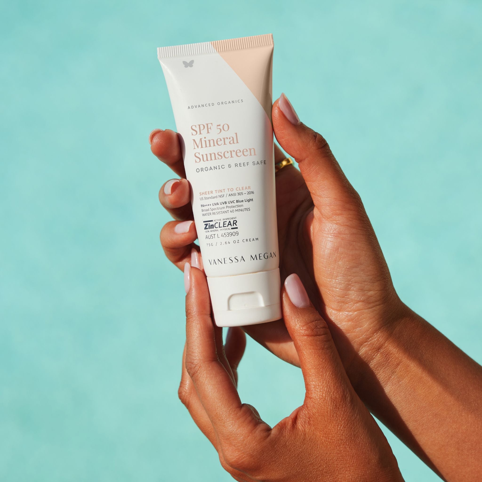 Hands holding the Vanessa Megan SPF 50 Mineral Sunscreen tube 75g, organic reef safe zinc sunscreen with broad spectrum UVA UVB protection in front of water. 