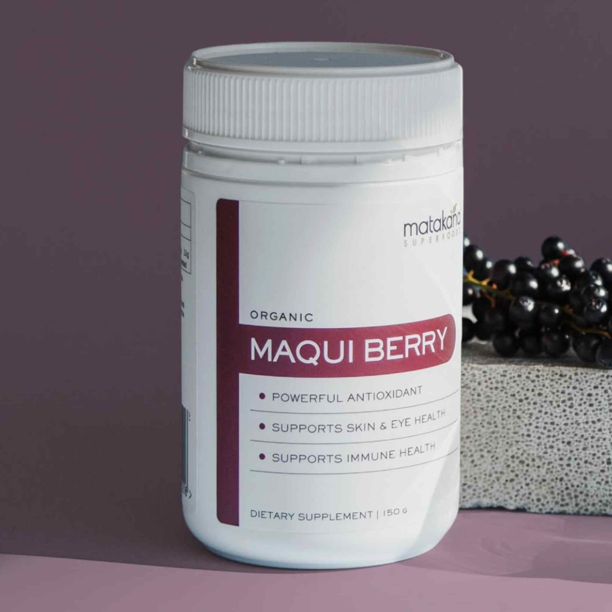 A white plastic bottle of Matakana Superfoods Organic Maqui Berry powder set against a dark purple background with a fresh bunch of maqui berries on the right.