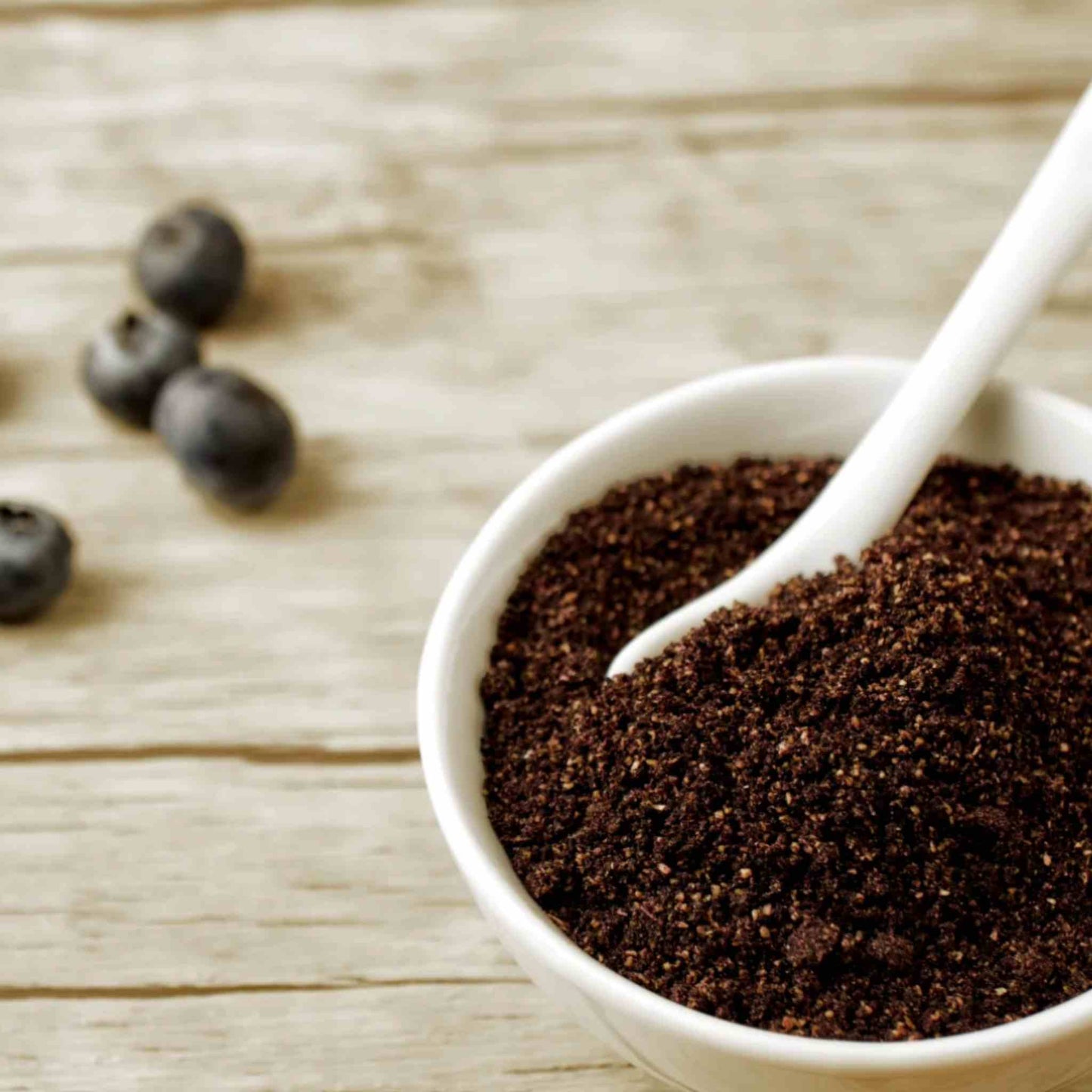 A white bowl filled with the Matakana organic Maqui berry powder, with a white spoon resting inside and a few berries on a light wooden surface in the background.