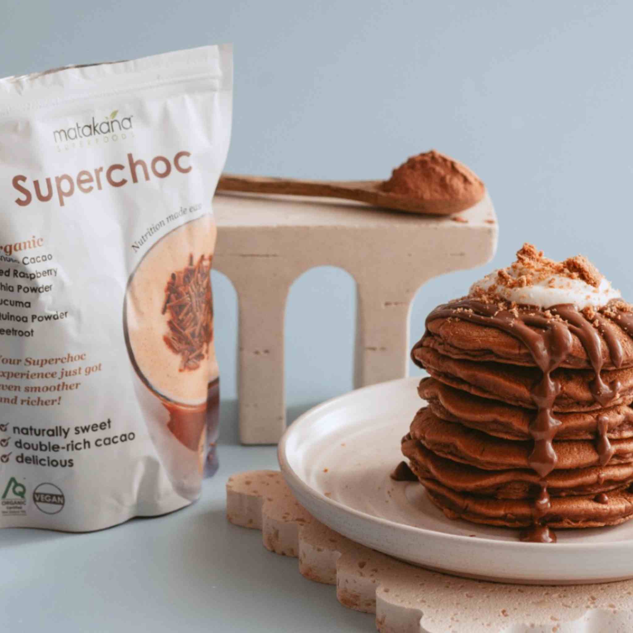 A bag of Matakana Superfoods Superchoc powder stands next to a tall stack of chocolate pancakes topped with coconut cream and drizzles on a white plate, with a wooden spoon holding Matakana Superfoods Superchoc powder on a small pedestal in the background.