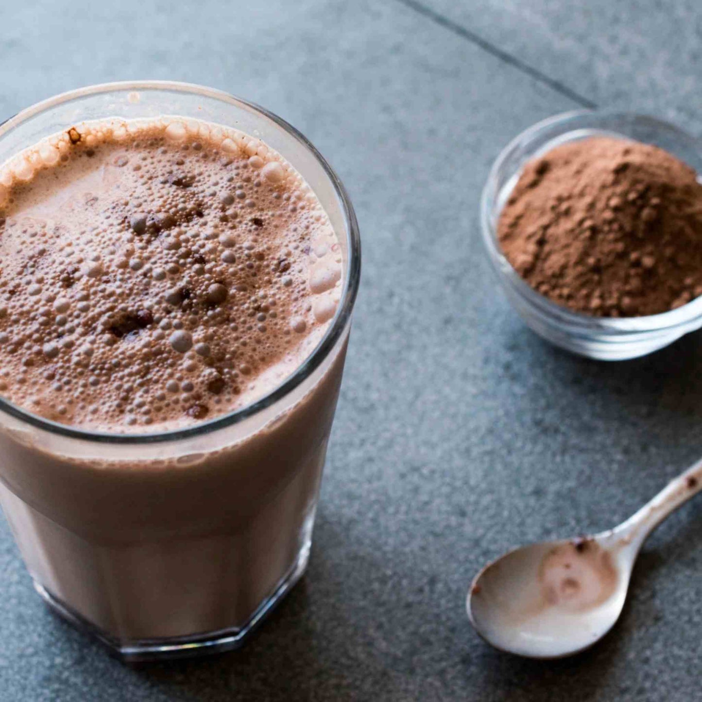A frothy of teh Matakana Superfoods Superchoc powder drink in a clear glass, with a small bowl of  the Superchoc powder and a spoon on a dark gray surface.