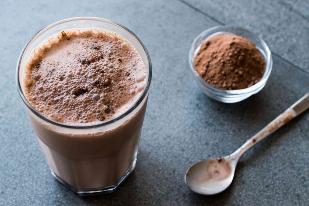 A glass of the Matakana Superfoods Superchoc shown on a dark gray surface next to a small bowl of brown powder and a spoon.