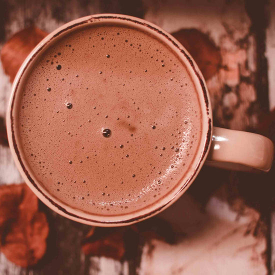 A top-down view of the Matakana Superfoods Superchoc drink in a beige ceramic mug, set against a rustic wooden background with warm autumn leaves.