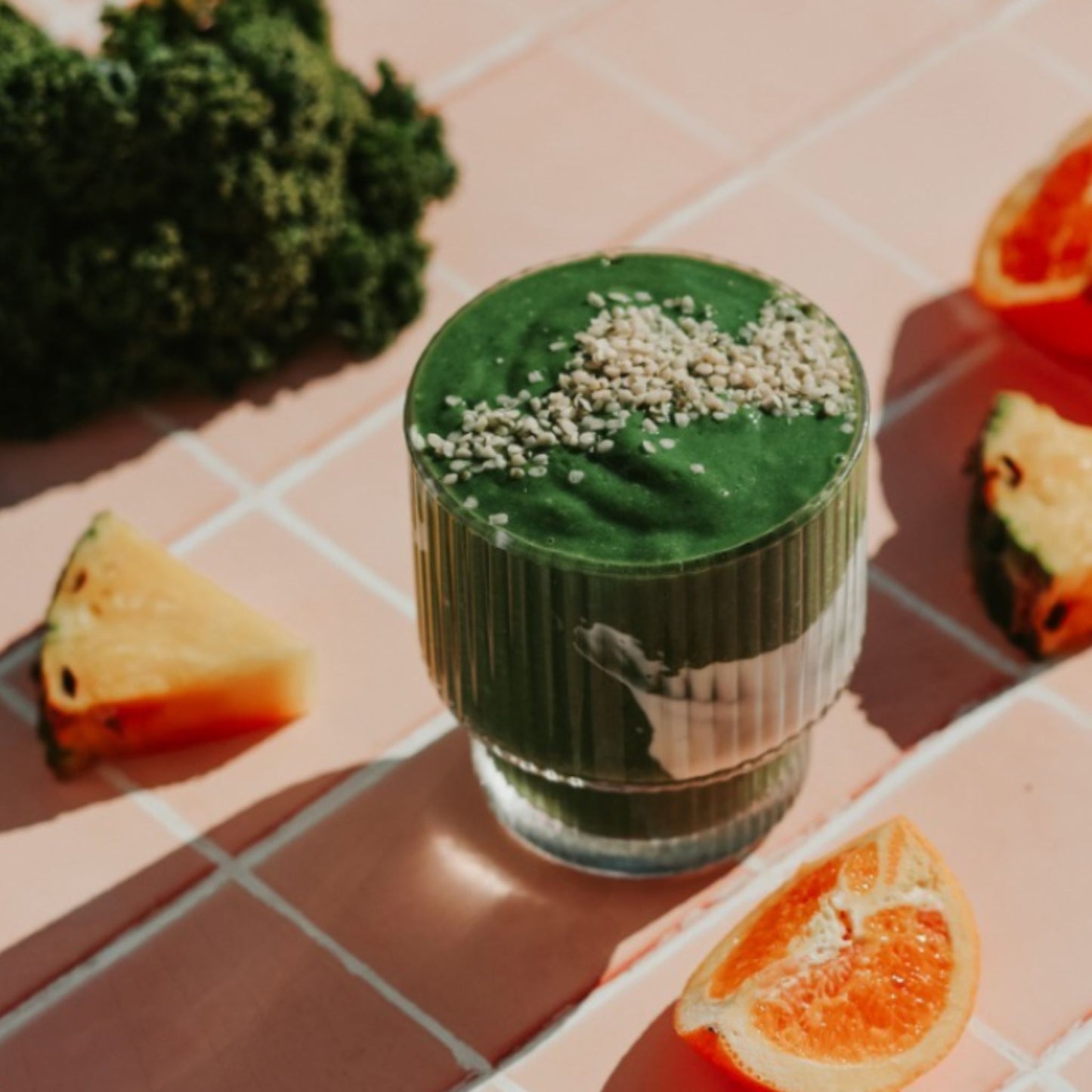 Healthy green smoothie  in glass topped with hemp seeds made with the  Matakana Superfoods Supergreens Powder shot next to fresh kale and cut fruit on sun lit surface. 
