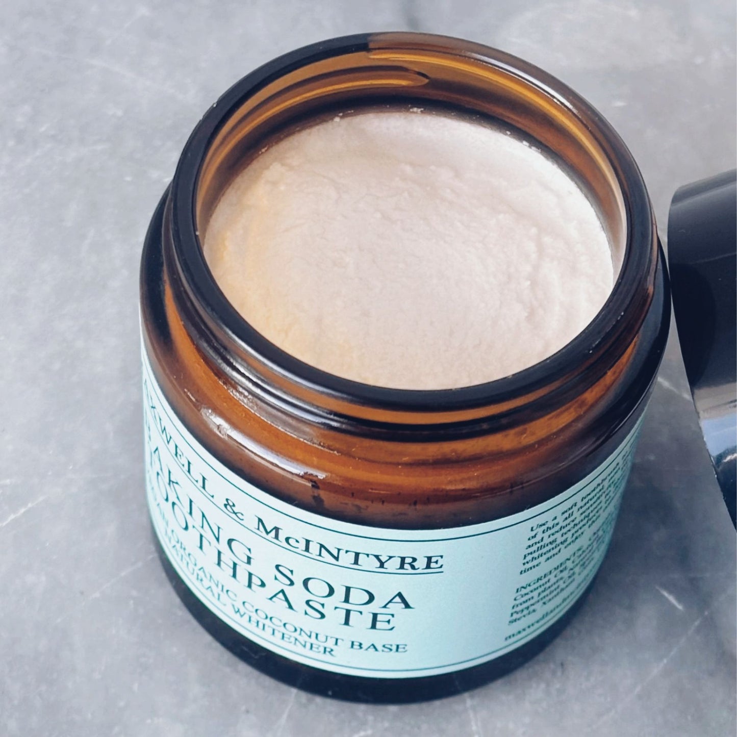 Close up of Maxwell & McIntyre Baking Soda Coconut Oil fluoride free toothpaste texture in amber glass jar with lid resting beside on marble surface.