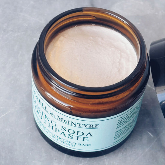Close up of Maxwell & McIntyre Baking Soda Coconut Oil fluoride free toothpaste texture in amber glass jar with lid resting beside on marble surface.