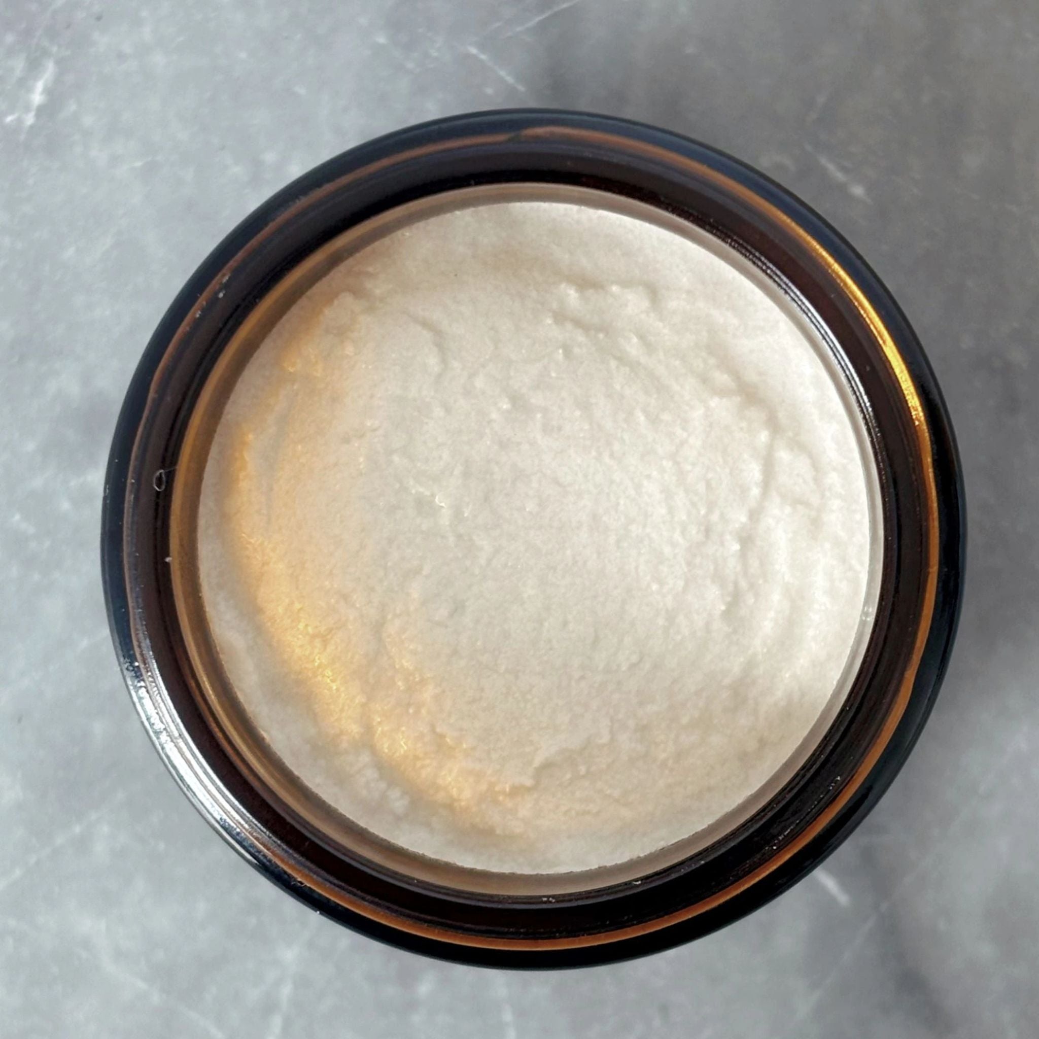 Top view of Maxwell & McIntyre Baking Soda Coconut Oil fluoride free toothpaste texture in amber glass jar on marble surface.