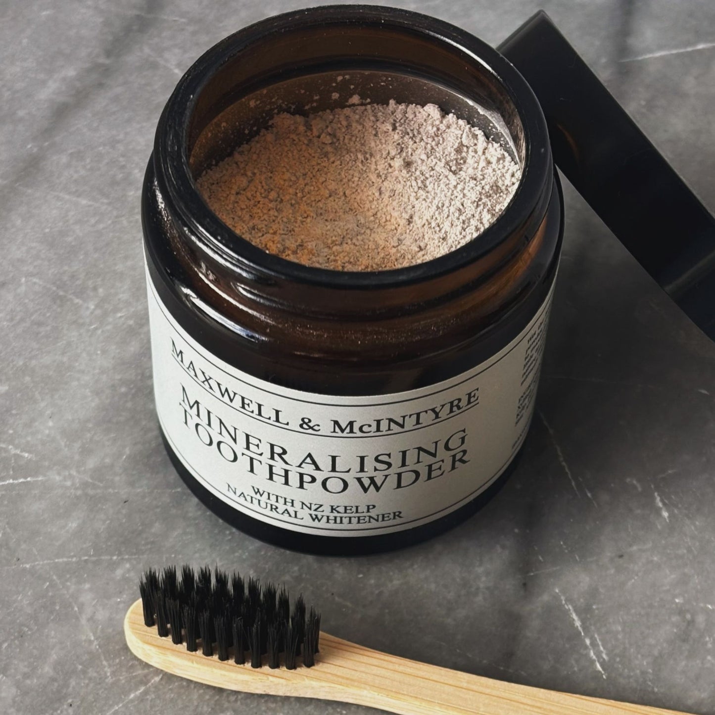Open amber glass jar of Maxwell and McIntyre Mineralising Toothpowder with NZ kelp beside black lid and CaliWoods bamboo toothbrush on grey stone surface.