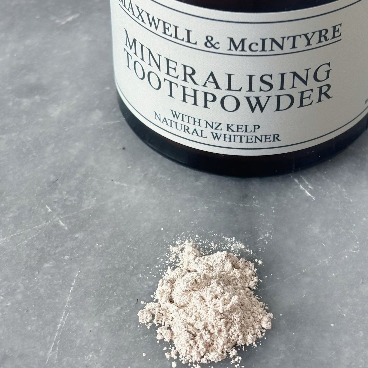 Maxwell and McIntyre Mineralising Toothpowder with NZ kelp natural whitener beside loose powder on stone surface.