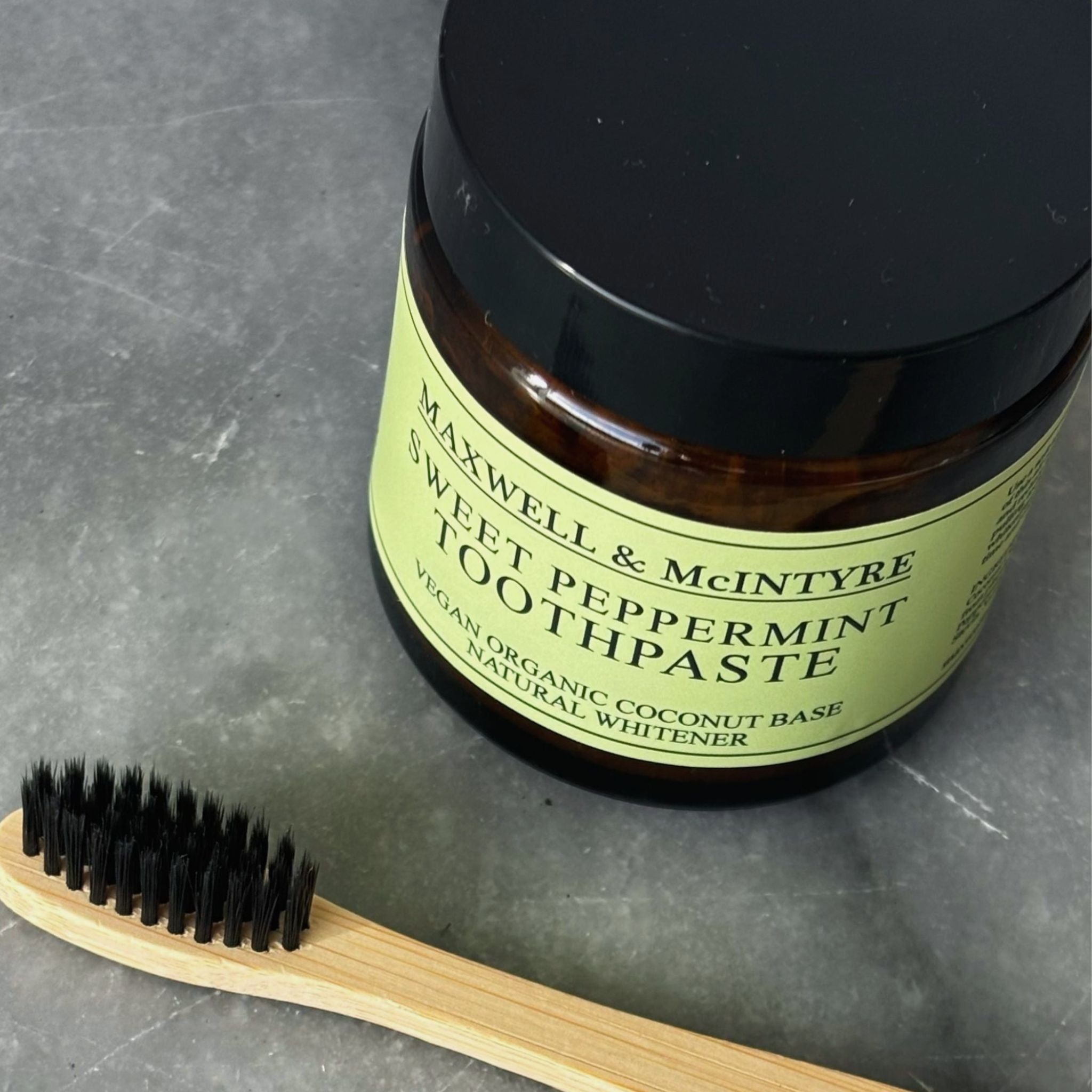 Maxwell & McIntyre Sweet Peppermint Toothpaste in amber glass jar with CaliWoods bamboo toothbrush on stone surface.