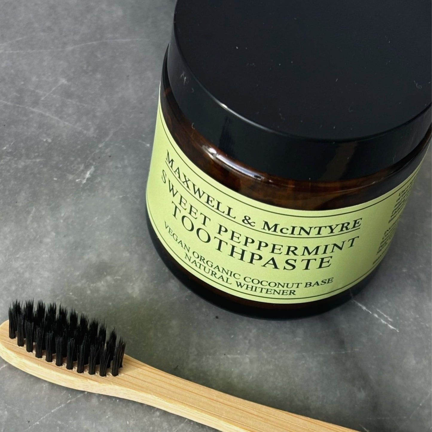 Maxwell & McIntyre Sweet Peppermint Toothpaste in amber glass jar with CaliWoods bamboo toothbrush on stone surface.