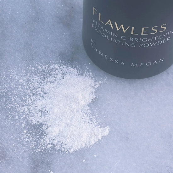 Close-up of the Metamorphosis Flawless Exfoliating Powder scattered on a light marble surface, showcasing its fine, powder texture beside the bottle.