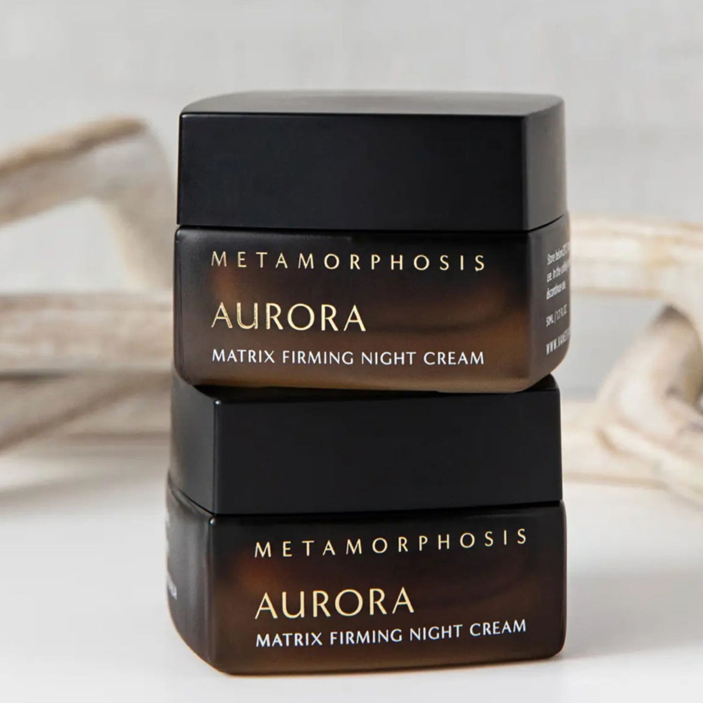 Two stacked jars of Metamorphosis Aurora Matrix Firming Night Cream, shown in dark brown and black packaging with gold lettering on a light, softly blurred background.