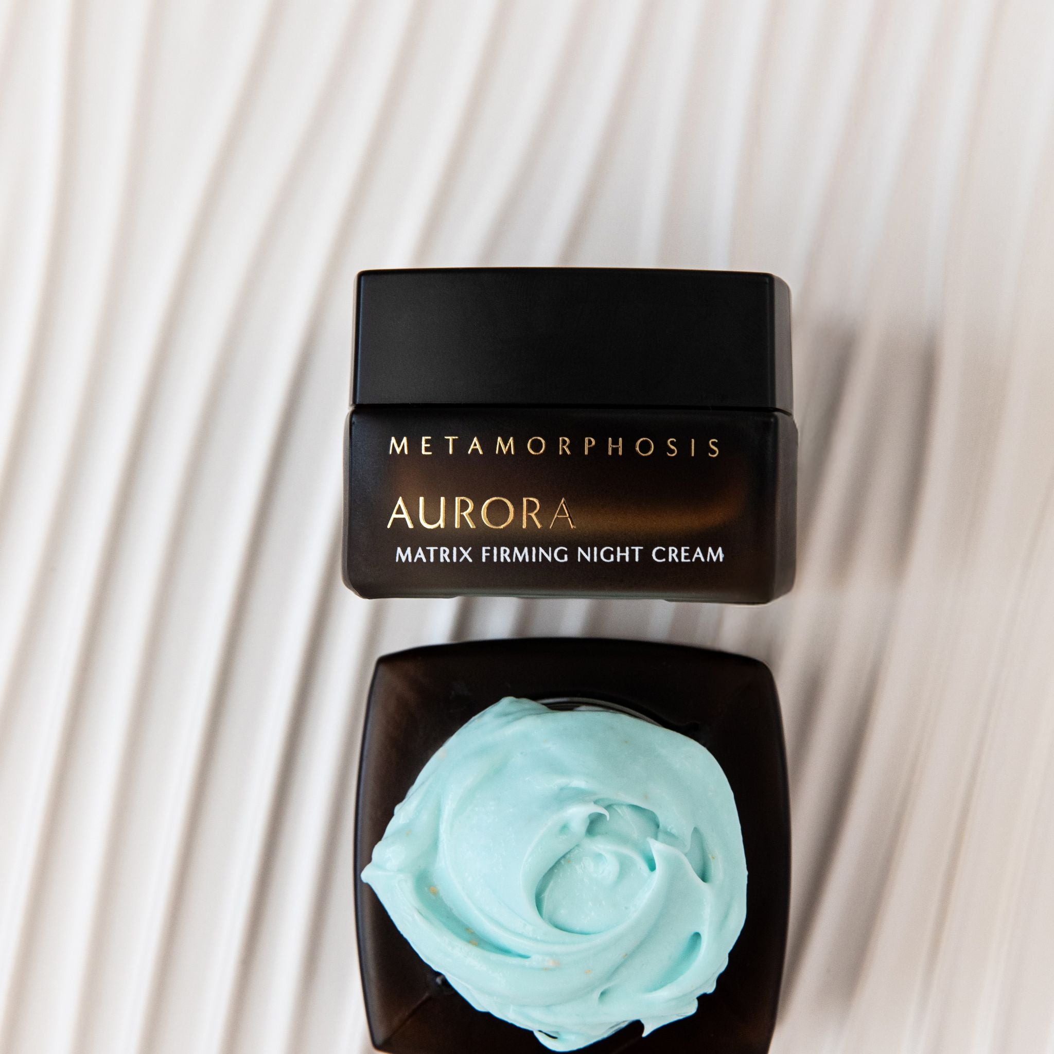 A jar of Metamorphosis Aurora Matrix Firming Night Cream shown closed with a black lid above, and another view of the open jar below displaying a swirl of pale mint blue cream, set against a cream-colored wavy background.