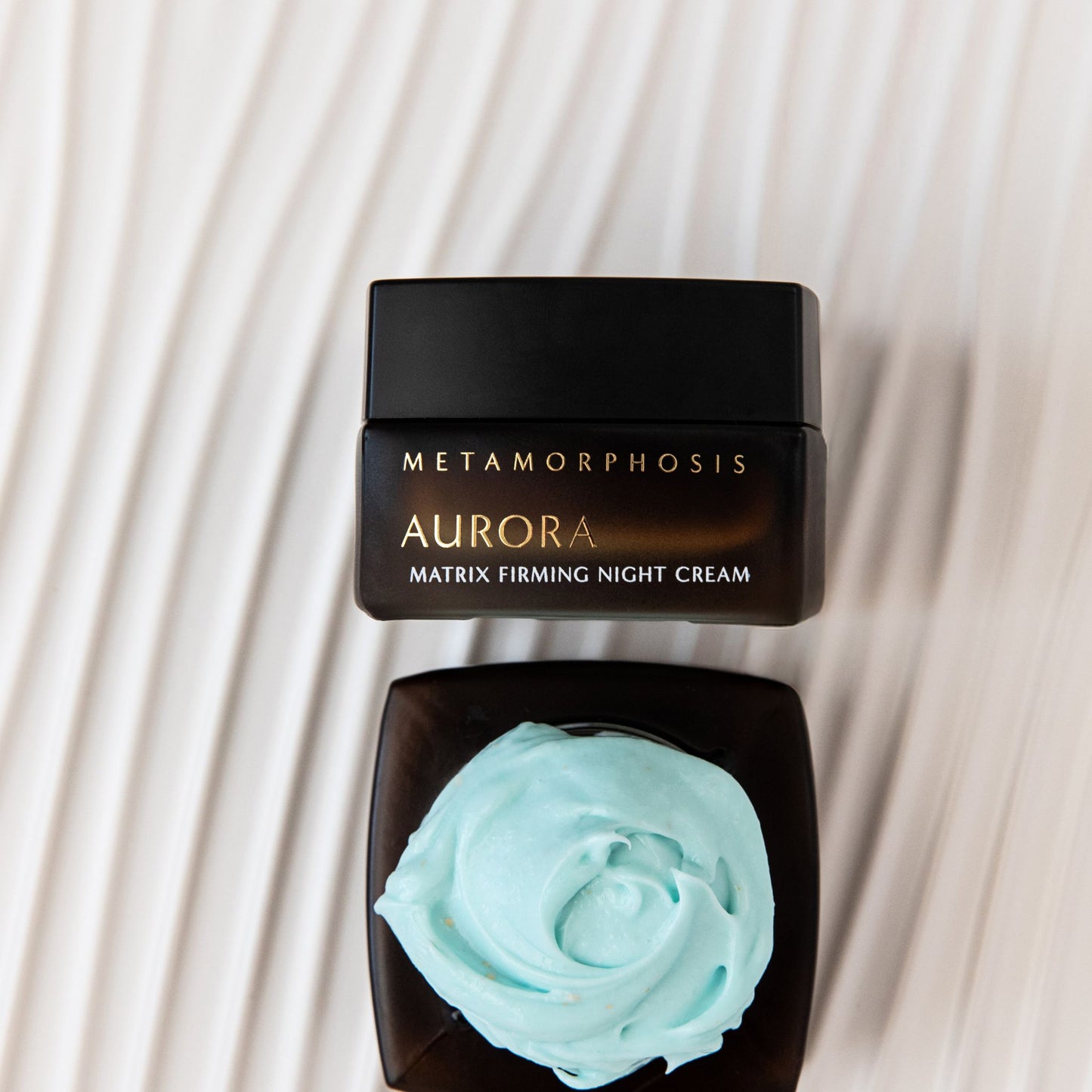A jar of Metamorphosis Aurora Matrix Firming Night Cream shown closed with a black lid above, and another view of the open jar below displaying a swirl of pale mint blue cream, set against a cream-colored wavy background.