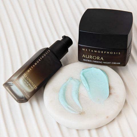 Metamorphosis Aurora Matrix Firming Night Cream shown with its black jar and The Illuminate Cellular Repair Day Cream dark pump bottle beside a white marble dish holding a swatche of each cream side by side, on a white ribbed surface.