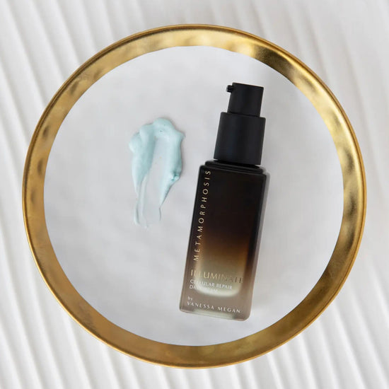 A bottle of VMetamorphosis Skincare Illuminate Cellular Repair Day Cream, placed on a white circular tray with a gold rim next to a smudge of Illuminate Cellular Repair Day Cream , on a white textured background.