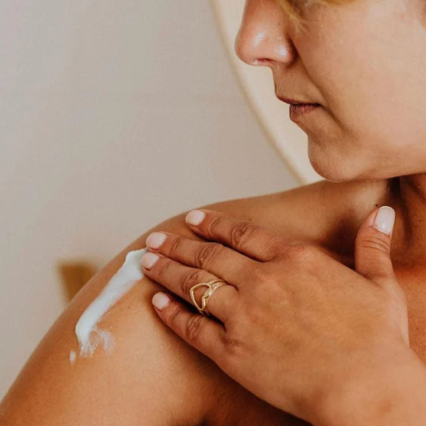 Close-up of a woman applying the Metamorphosis Skincare Wild Yam Concentrate Dermal Cream  to her shoulder while wearing a gold heart-shaped ring on her finger.