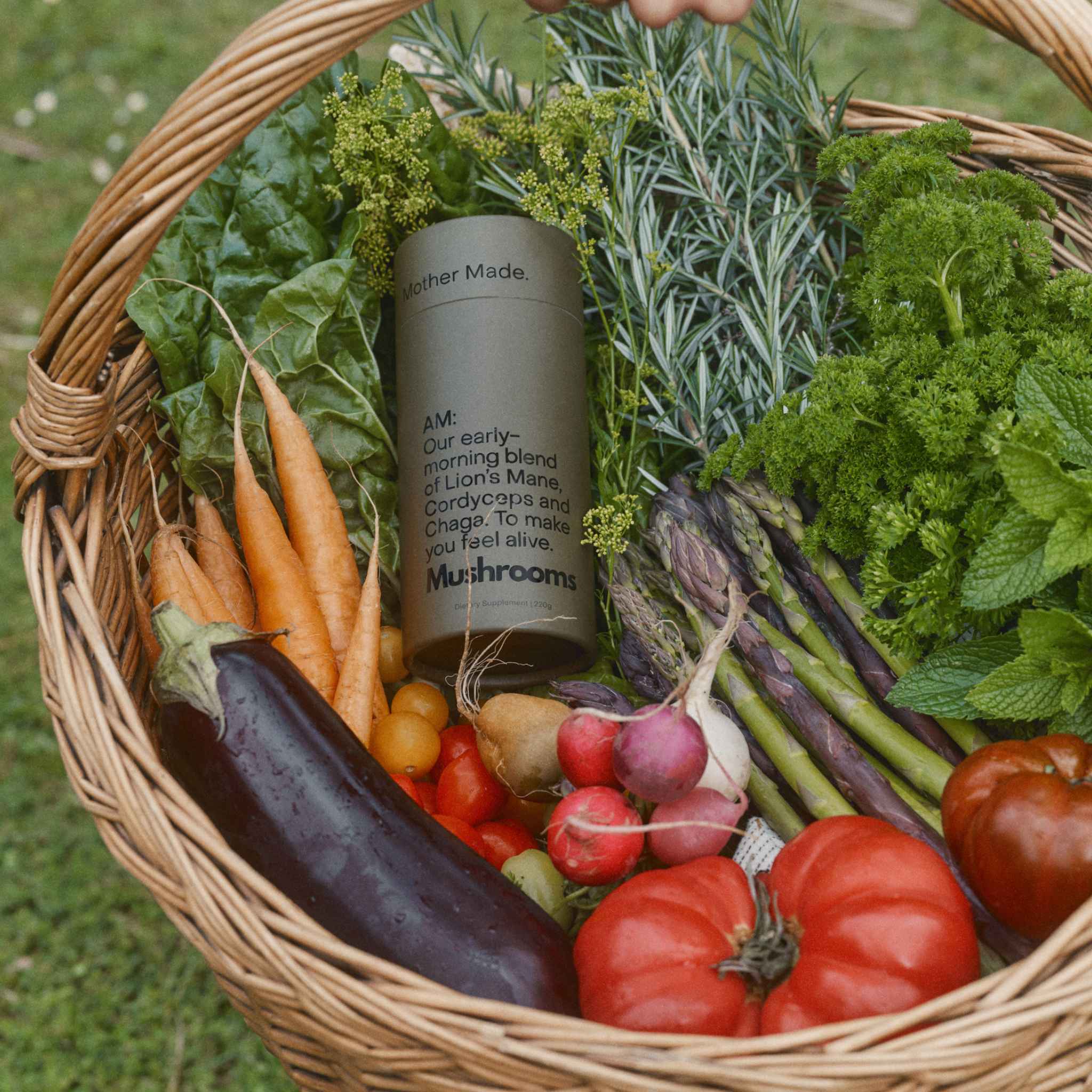 A wicker basket filled with fresh vegetables and herbs, including carrots, eggplant, tomatoes, radishes, asparagus, parsley, and rosemary, with a  canister of the Mother Made AM Blend Morning mushroom powder placed in the center.