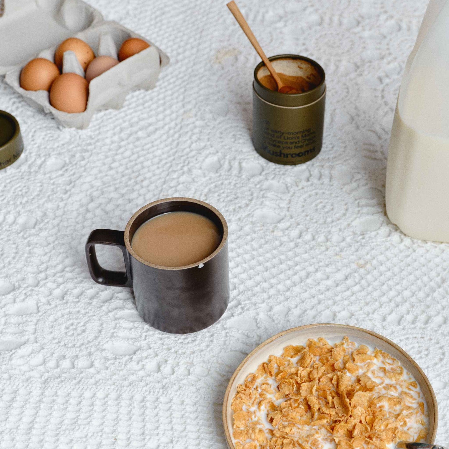 A  breakfast scene on a white textured blanket with a dark mug filled with coffee made with the Mother Made AM Blend, a bowl of cereal with milk, eggs in a carton, and a small jar of honey with a spoon.