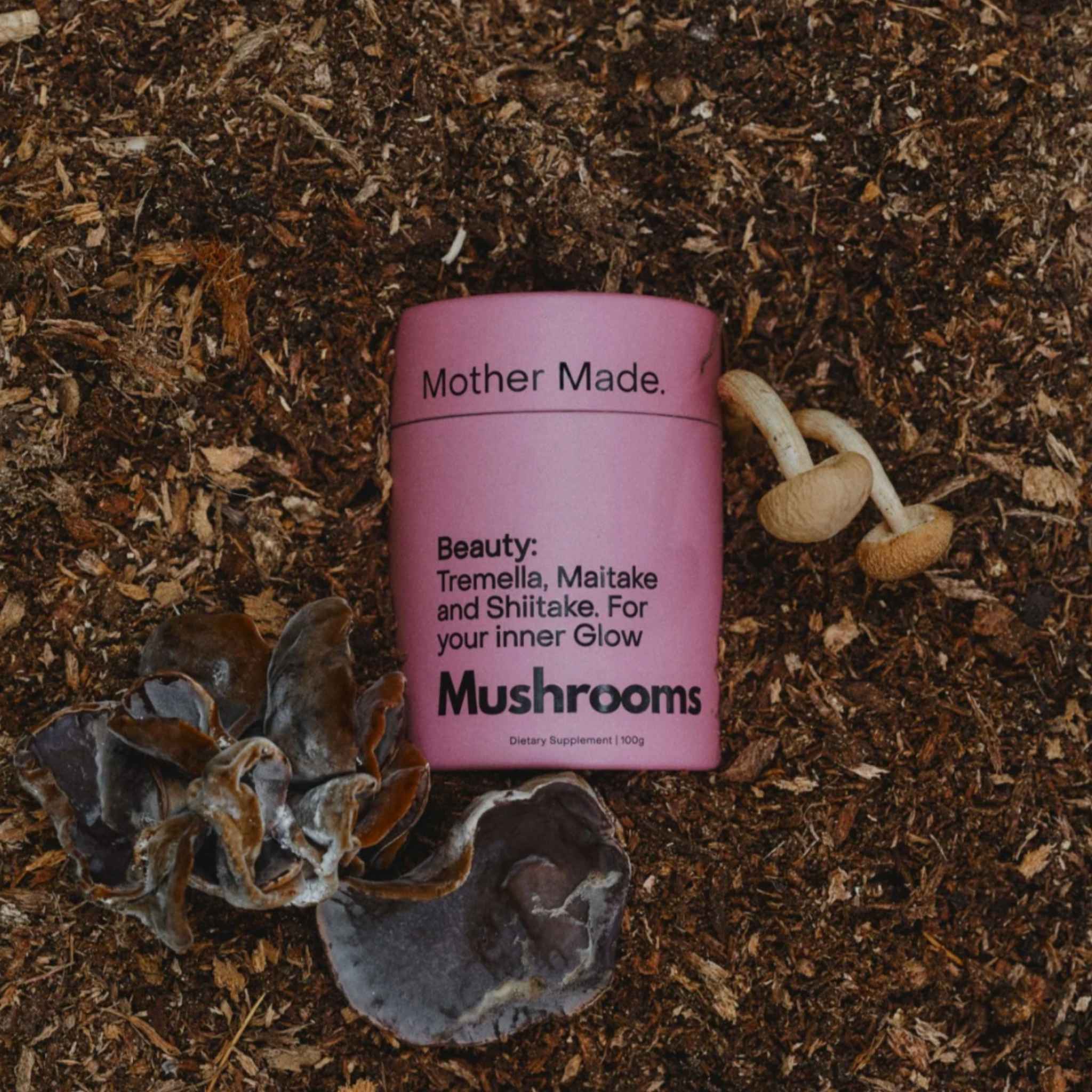 A pink cylindrical tin of the Mother Made. Beauty: Tremella, Maitake and Shiitake blend nestled in the earth with fungi around it.