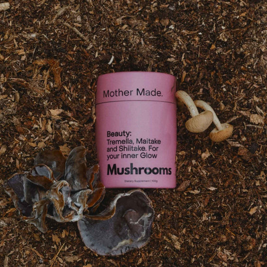 A pink cylindrical tin of the Mother Made. Beauty: Tremella, Maitake and Shiitake blend nestled in the earth with fungi around it.
