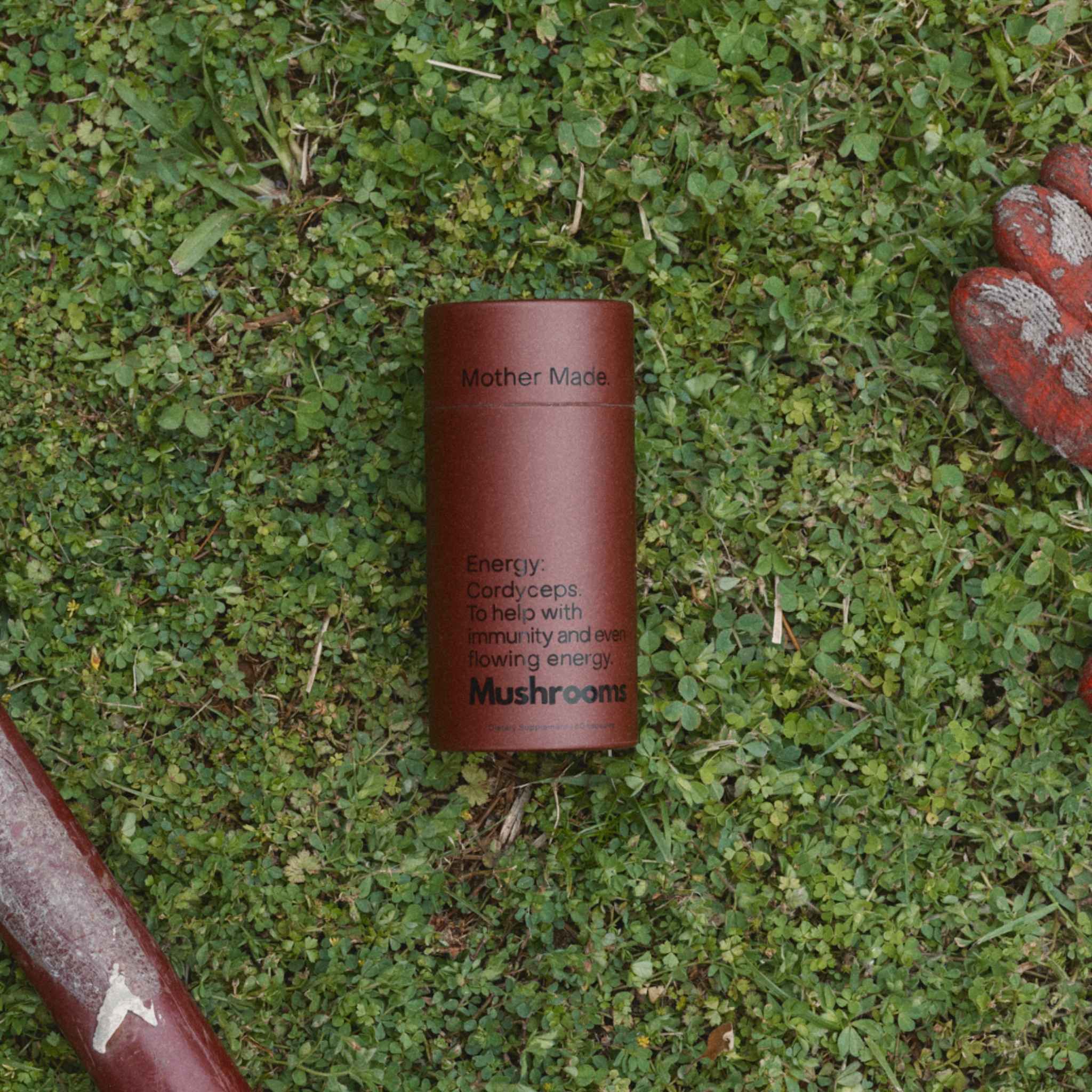 A canister of the Mother Made Mushrooms Energy placed on green grass, with gardening gloves partially visible in the corners.