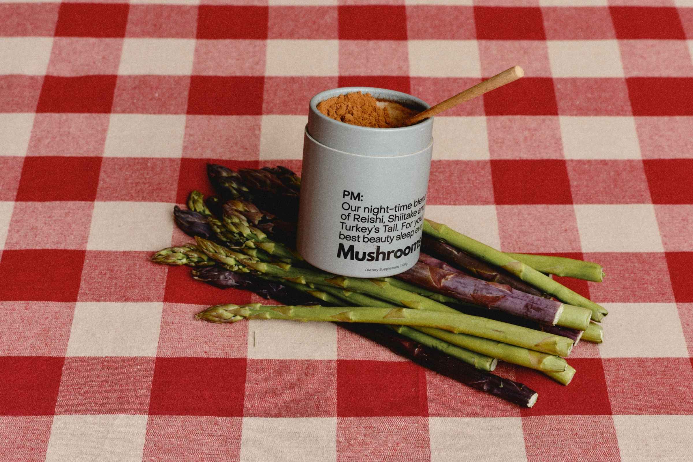A 100g cardboard canister of the Mother Made PM mushrrom powder Blend with a wooden spoon, placed on a red and white checkered tablecloth on top of a bunch of green and purple asparagus spears.