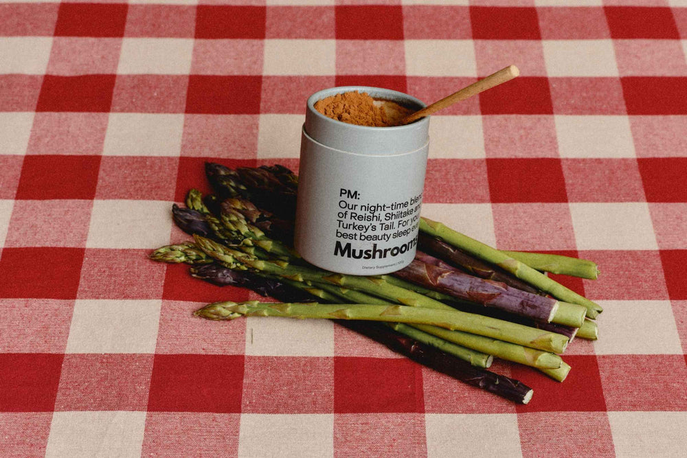 A 100g cardboard canister of the Mother Made PM mushrrom powder Blend with a wooden spoon, placed on a red and white checkered tablecloth on top of a bunch of green and purple asparagus spears.