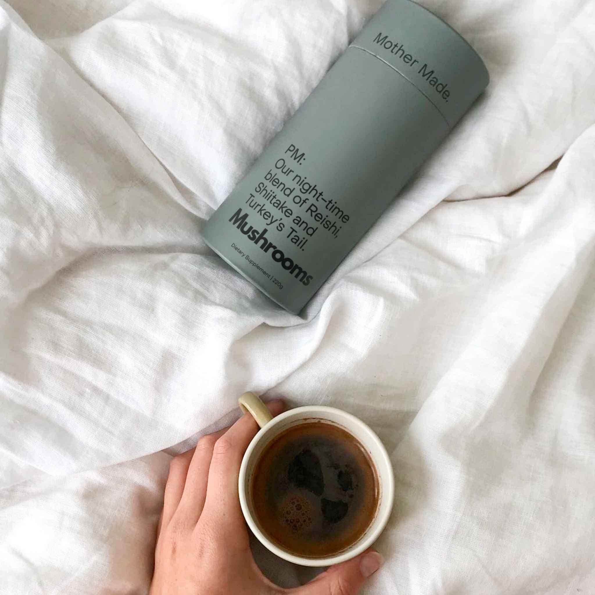 A hand holds a white ceramic mug filled with a warm drink of the mother made pm on rumpled white bedding, next to a 220g cardboard canister of the Mother Made PM Blend.