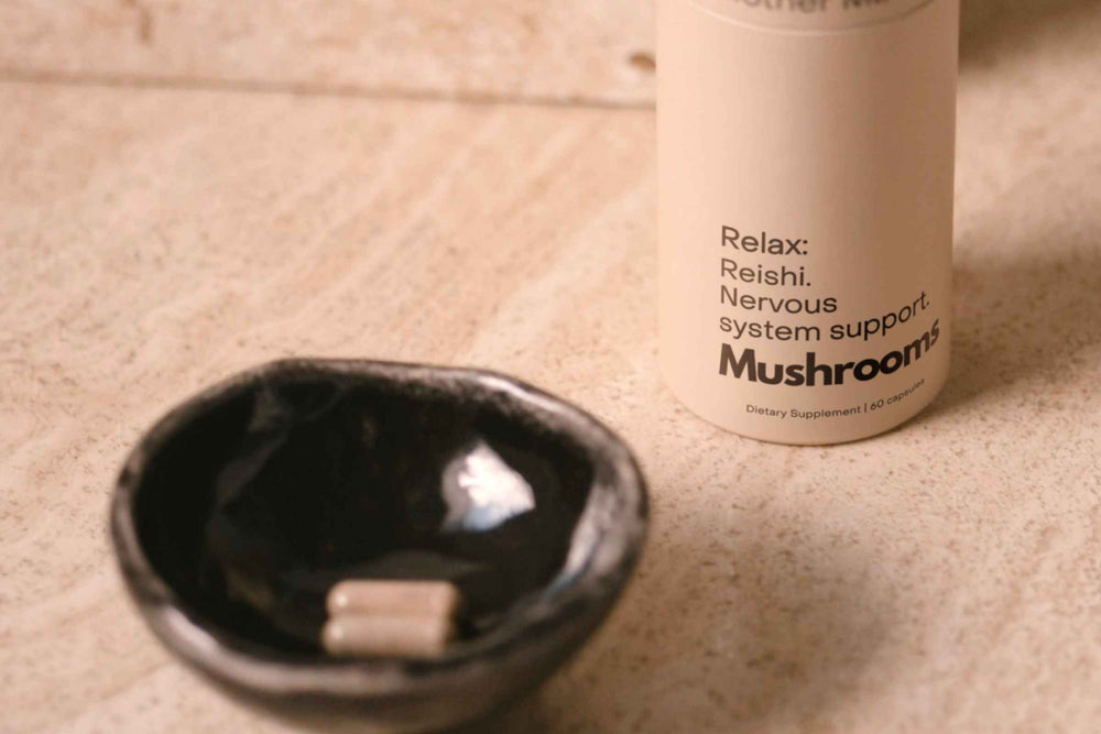 A close-up of  the 'Mushrooms Relax: Reishi, nervous system support mushroom capsules  next to a small dark ceramic dish containing a  couple of Reishi capsules on a textured beige surface.