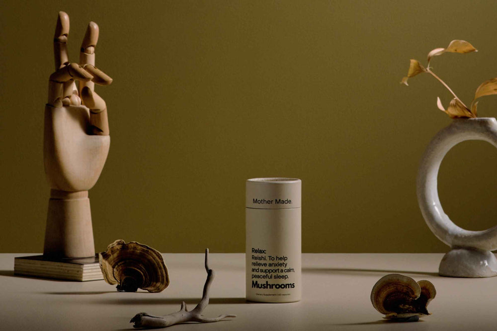 Mother Made Relax Reishi Capsules stands on a tabletop against a warm olive-green background, styled with a wooden hand sculpture, shell-like decor, Reishi  mushrooms and a white ceramic ring vase.