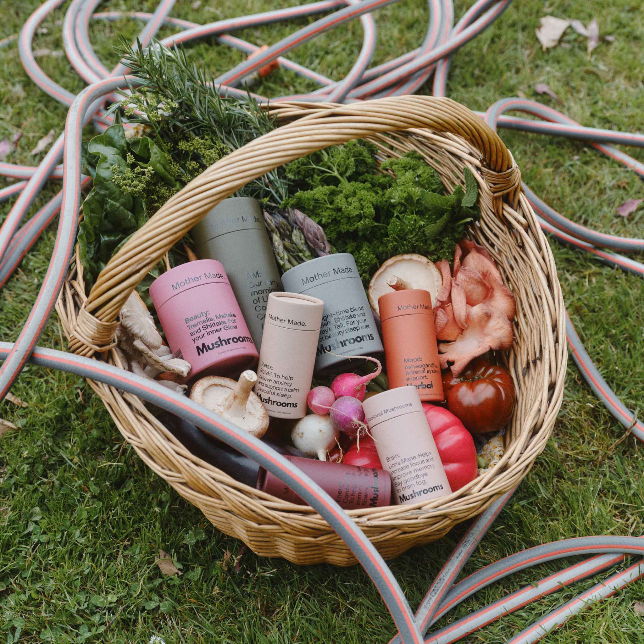 A wicker basket filled with The Mother Made product rane of medicinal mushroom powders and capsules and ashwagandha mood caps along with fresh greens, mushrooms, radishes, and tomatoes, set on a grassy lawn with garden hoses in the background.