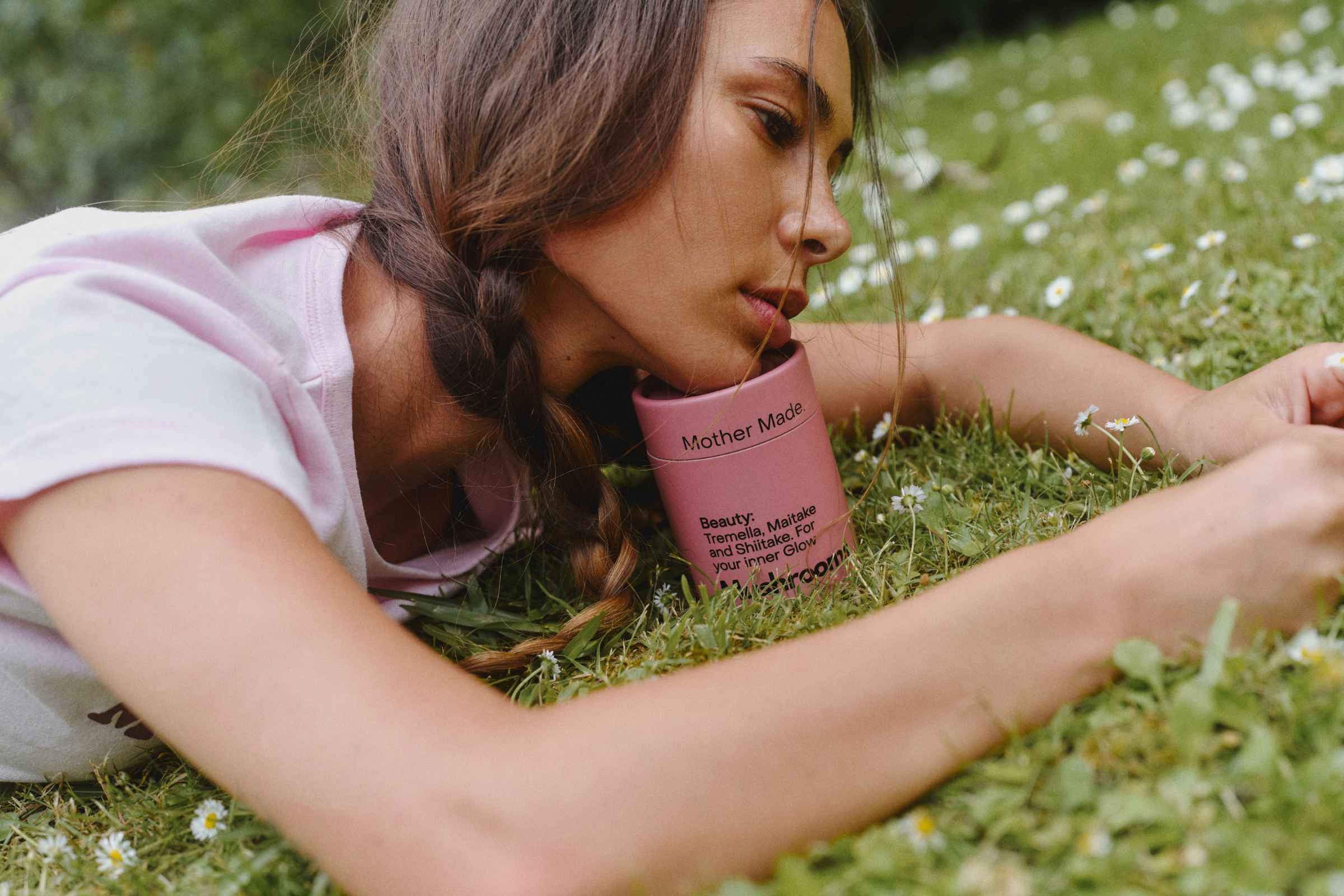 A woman lying on green grass among small white flowers, resting her face on a canister of the Mother Mother Beauty Blend.