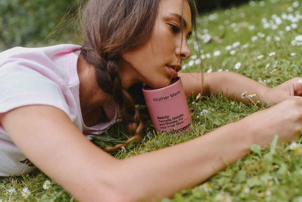 A woman lying on green grass among small white flowers, resting her face on a canister of the Mother Mother Beauty Blend.