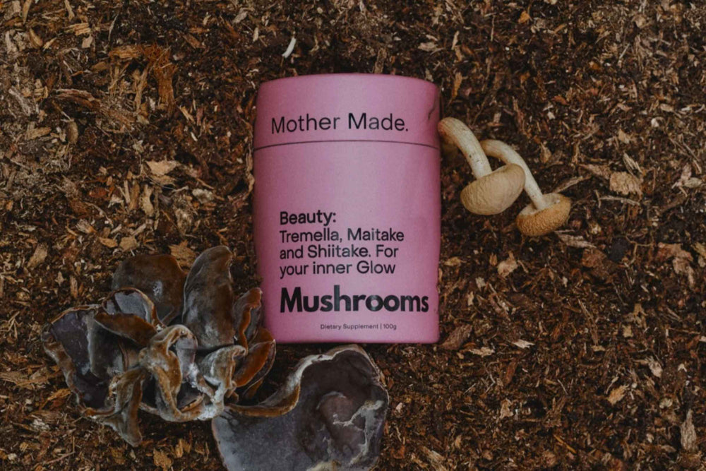 A pink cylindrical tin of the 'Mother Made. Beauty blend : Tremella, Maitake and Shiltake nestled in earth with Maitake and Shiltake beside it. 