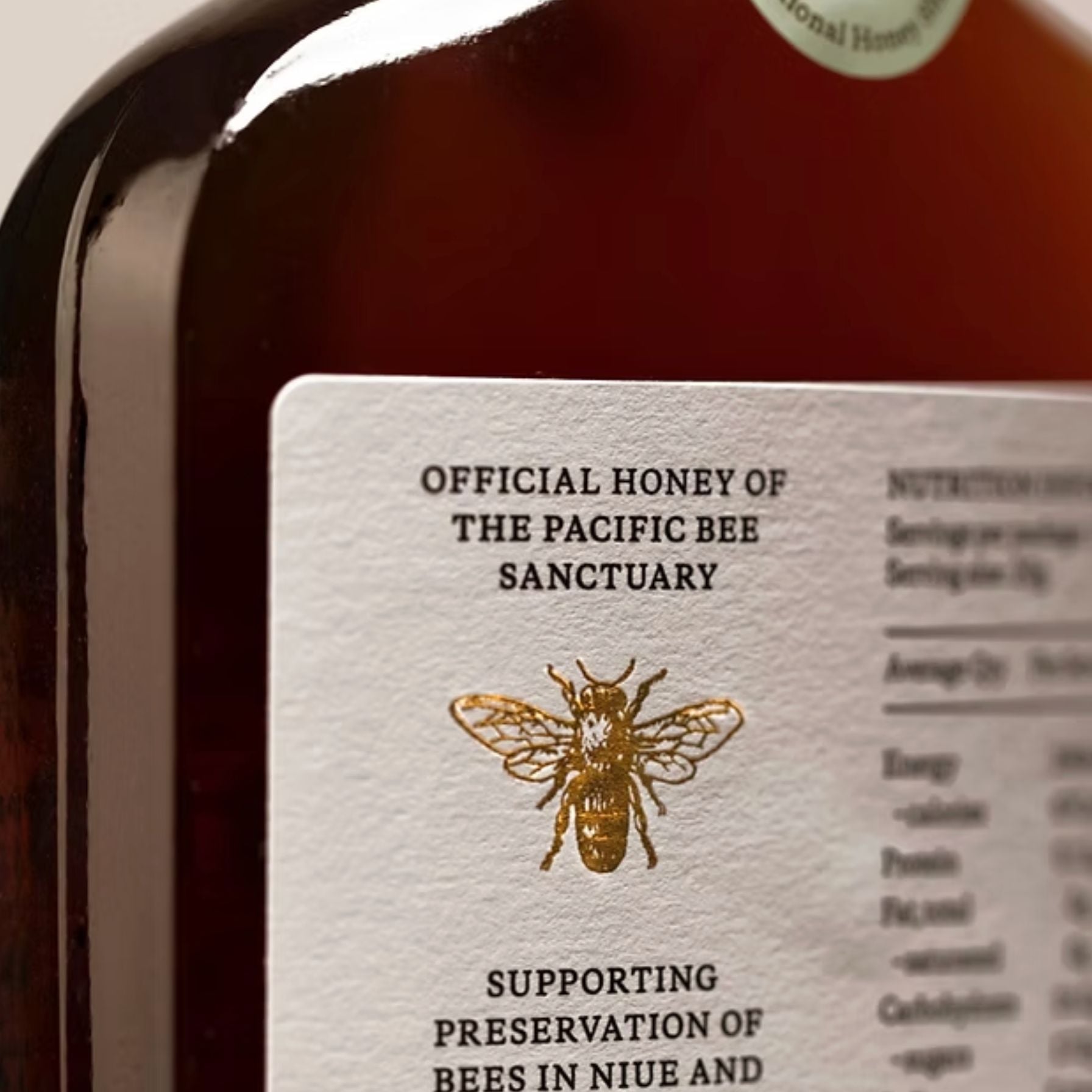 Close-up of the back of the Niue Honey bottle showcaseing its dark amber hue with a white label reading 'Official Honey of the Pacific Bee Sanctuary' and a gold bee illustration.