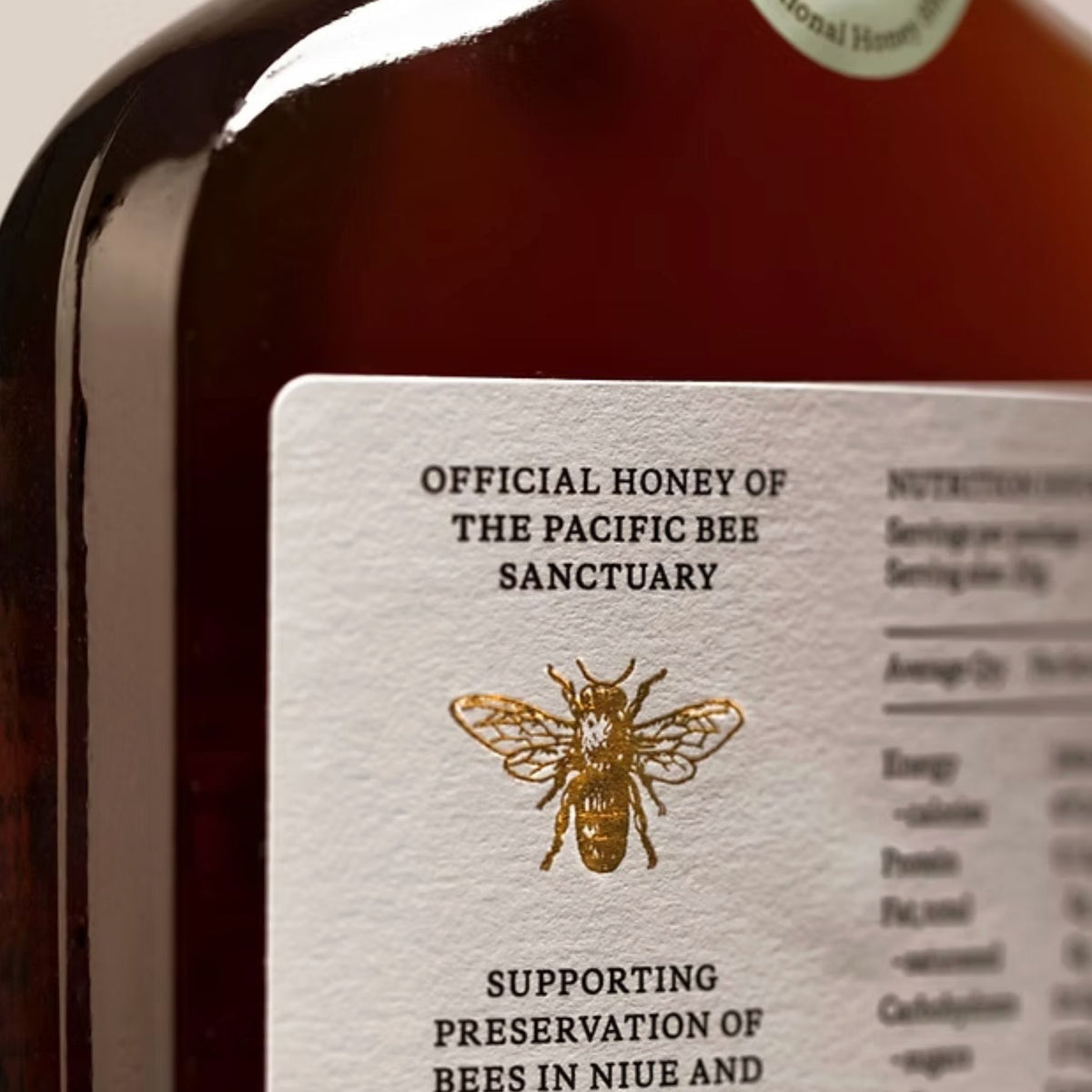 Close-up of the back of the Niue Honey bottle showcaseing its dark amber hue with a white label reading 'Official Honey of the Pacific Bee Sanctuary' and a gold bee illustration.